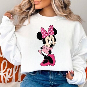 Puede incluir: Sudadera blanca con una imagen de dibujos animados de Minnie Mouse con un vestido rosa y un lazo rosa con lunares blancos.