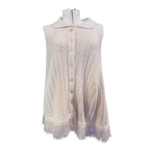 May include: Cream-colored, button-down knit poncho with a collar and fringe trim. The poncho features a cable knit design and is displayed on a mannequin. The garment is sleeveless and has a flowing, asymmetrical hemline.