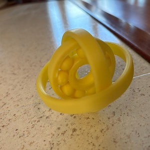 Puede incluir: Un juguete de plástico amarillo con una esfera de bolas amarillas más pequeñas en el centro. El juguete está hecho de anillos entrelazados.