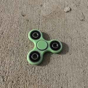 May include: A green fidget spinner with three black bearings. The spinner is on a grey concrete surface.