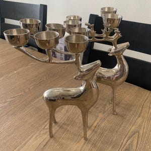May include: Two silver-toned metallic reindeer figurines, each featuring multiple candle holders integrated into their antlers. The polished, reflective surfaces of the deer bodies and the cup-shaped candle holders create a unified, sleek appearance.