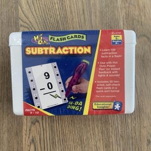 May include: A white plastic box containing Hot Dots Subtraction flash cards. The packaging features a blue and red design with text including 'SUBTRACTION', 'Learn 100 subtraction facts in a flash', and 'Includes 50 two-sided, self-check flash cards'.