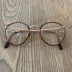 May include: Round eyeglasses featuring a tortoiseshell pattern on the rims and thin gold-tone metal temples. The bridge is also gold-tone metal, and the text Giorgio Armani is printed on the upper part of the left lens frame in small letters.