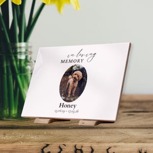 Op de afbeelding: Witte keramische gedenkplaat met de tekst "in loving memory" en een ovale foto van een hond genaamd Honey. De plaat wordt ondersteund door een houten standaard en bevat de data 23.04.19 - 17.03.26.