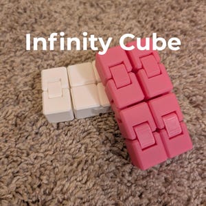 May include: Two infinity cubes, one white and one pink, are displayed on a textured, neutral-toned surface. The text "Infinity Cube" is written in white across the top of the image. The cubes are designed with multiple connected blocks.