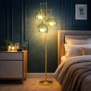 May include: A gold-colored floor lamp with three green glass shades. The lamp has a round base and a tall, slender stem. The shades are a light green color and have a ribbed design. The lamp is in a bedroom setting.