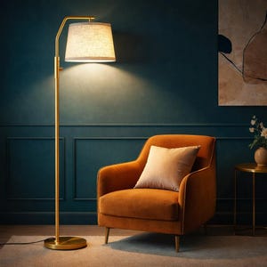 May include: A gold floor lamp with a beige lampshade illuminates an orange armchair with a beige pillow. The lamp has a curved arm and a round base. The background is a dark teal wall with a painting and a small side table.
