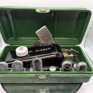 May include: A black Singer buttonholer attachment with a white dial featuring a red S, housed in a textured green plastic case. The set includes several grey metal attachments arranged in slots within the base of the open container.