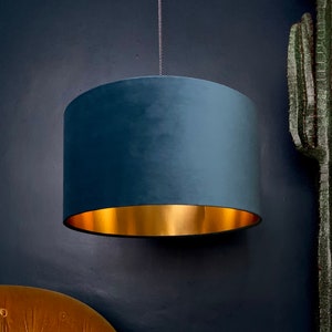 Op de afbeelding: Een lampenkap van tealblauwe fluweel met een gouden rand. De lampenkap hangt aan een plafondarmatuur.