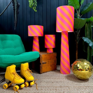 May include: Three striped lamps with pink and orange stripes stand on a wooden floor in front of a dark green armchair. A disco ball sits on the floor in front of the tallest lamp. Two yellow roller skates are on the floor in front of the armchair.
