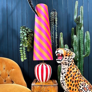 May include: A pink and orange striped lampshade with a white and red striped ceramic base. The lamp is sitting on a wooden table next to a ceramic leopard statue.