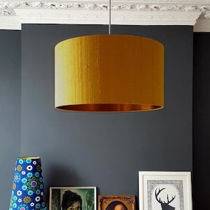 May include: A gold drum lampshade with a textured fabric finish hangs from the ceiling in a room with dark gray walls. The lampshade is illuminated and casts a warm glow. There are framed pictures and a table lamp with a blue and white floral patterned shade on a wooden table below the lampshade.