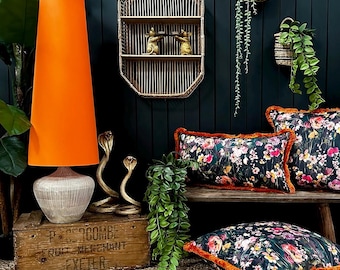 May include: A tall floor lamp with a bright orange conical shade and a ribbed glass base, sitting on a wooden crate. Nearby are two gold-colored cobra figurines, a rectangular floral pillow with orange fringe, and a square floral pillow with orange fringe.