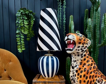 Op de afbeelding: Een zwart-wit gestreepte lampenkap met een blauw-wit gestreepte keramische basis. De lamp staat op een houten kist met de tekst "DUCTS LTD" en "N 474". Op de achtergrond staat een keramiek leeuwenbeeld.