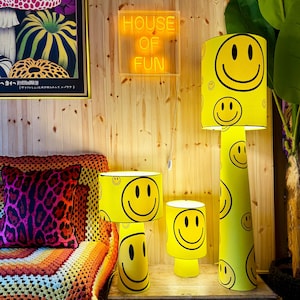 May include: A collection of yellow lamps with black smiley faces. The lamps are different sizes, including a floor lamp and two table lamps. The background includes a wooden wall, a neon sign that says "HOUSE OF FUN", and a colorful couch.