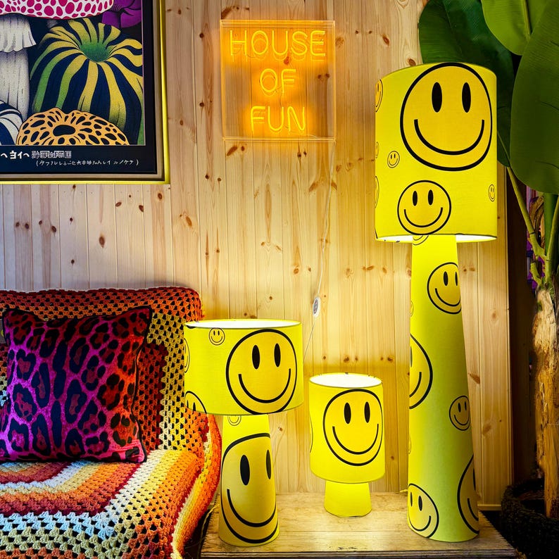 May include: A collection of yellow lamps with black smiley faces. The lamps are different sizes, including a floor lamp and two table lamps. The background includes a wooden wall, a neon sign that says "HOUSE OF FUN", and a colorful couch.