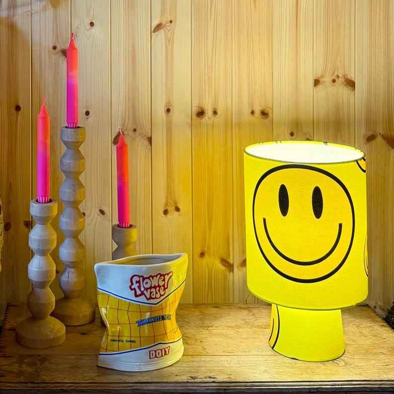 May include: A yellow smiley face lamp, three wooden candle holders with pink and orange candles, and a ceramic flower vase shaped like a snack bag are displayed on a wooden surface. The vase has the words "Flower Vase" and "DOIY" printed on it.