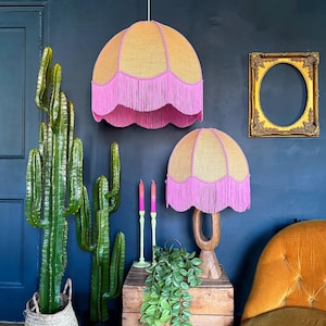 May include: Two decorative lampshades with a scalloped edge and pink fringe. The lampshades are gold and pink, with one hanging from the ceiling and the other on a wooden base. A large green cactus and a gold framed oval mirror are also in the image.