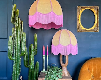 May include: Two decorative lampshades with a scalloped edge and pink fringe. The lampshades are gold and pink, with one hanging from the ceiling and the other on a wooden base. A large green cactus and a gold framed oval mirror are also in the image.