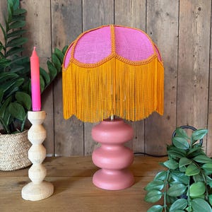 Puede incluir: Una lámpara de mesa rosa con una base escalonada y una pantalla rosa brillante. La pantalla tiene un ribete naranja y largas borlas naranjas. Una vela rosa y un candelabro de madera están a la izquierda.