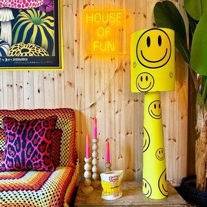May include: A yellow floor lamp with black smiley faces, a neon "HOUSE OF FUN" sign, and a leopard print pillow. The lamp has a cylindrical base and a lampshade with a large smiley face.