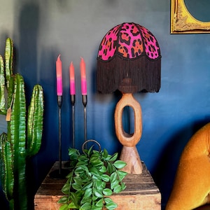 Può includere: Una lampada da tavolo in legno con paralume con stampa leopardata rosa e arancione e frange nere. Tre candele sfumate rosa e arancioni in portacandele in metallo nero. Un cactus verde e una pianta ricadente sono anche sul tavolo.