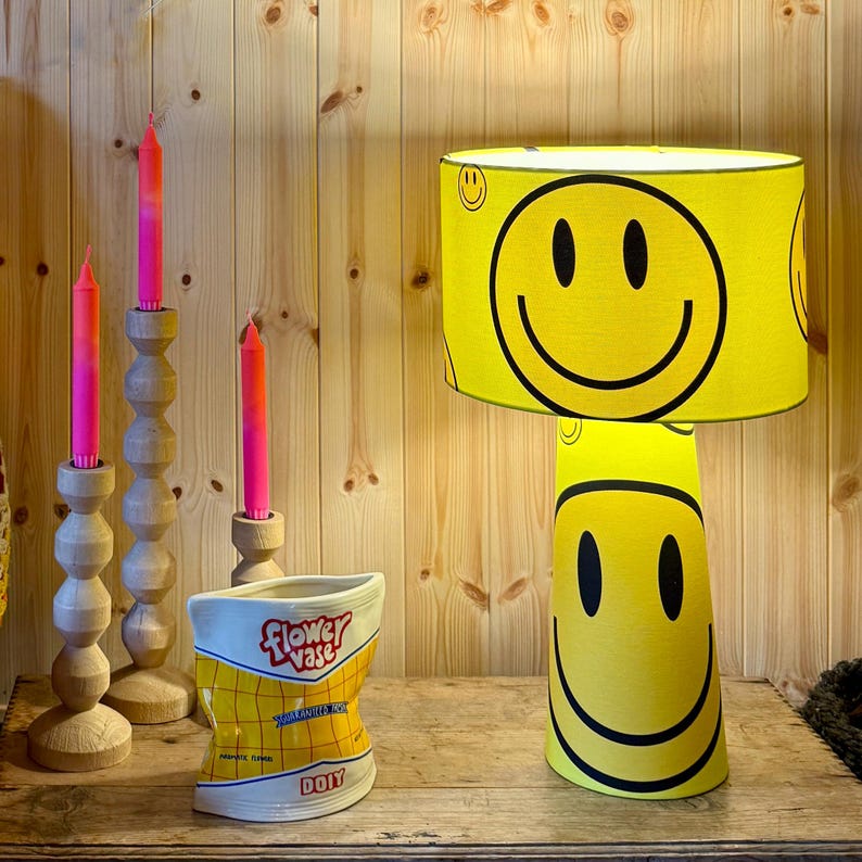 May include: A yellow lamp with a smiley face design, a ceramic flower vase, and three wooden candle holders with pink candles. The lamp has a cylindrical shade and a base with a large smiley face. The vase is shaped like a bag of chips.