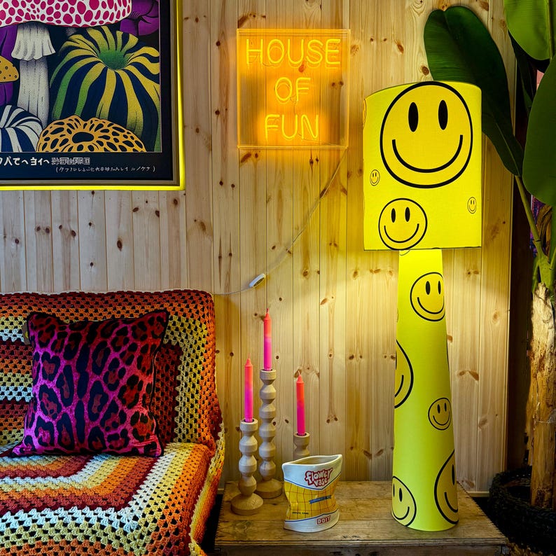 May include: A yellow lampshade with a smiley face design, a neon sign that says "HOUSE OF FUN", and a decorative pillow with a pink leopard print pattern. The room has a wooden wall and a colorful blanket.