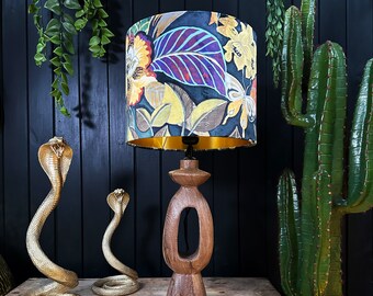 Op de afbeelding: Een houten tafellamp met een donkerhouten voet en een zwarte lampenkap met een kleurrijk bloemenpatroon. De lampenkap heeft een gouden rand. De lamp staat op een houten tafel met twee gouden slangfiguren.