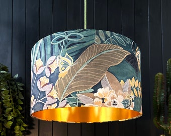 Op de afbeelding: Een gouden en groene lampenkap met een tropisch bloemenpatroon. De lampenkap heeft een gouden rand en toont een groot blad, bloemen en vogels.