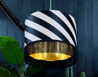 May include: A black and white striped lampshade with a gold interior and black fringe. The lampshade is hanging from a green cord against a dark blue wall. Decorative lighting for a living room or bedroom.