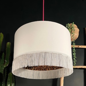 Può includere: Paralume color crema con frange sul fondo e interno con stampa leopardata. Appeso a un cavo rosa. Il design combina elementi moderni e bohémien.