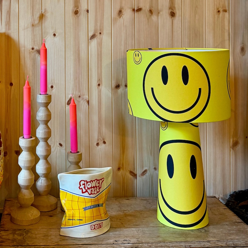 May include: A yellow smiley face lamp with a matching lampshade, a ceramic vase shaped like a bag of chips, and three wooden candle holders with pink and orange candles. The vase reads "Flower Vase" and "DOY".