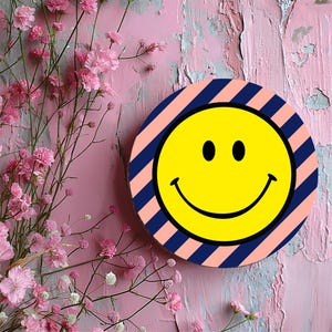 Peut inclure: Un dessous de verre rond avec un motif rayé rose et bleu. Un smiley jaune se trouve au centre du dessous de verre.