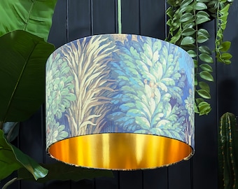 Op de afbeelding: Een cilindervormige lampenkap met een botanische print in blauw, groen en beige tinten. De binnenkant is gevoerd met glanzend goudkleurig materiaal. De lampenkap hangt aan een groen snoer, wat een decoratieve verlichting creëert.