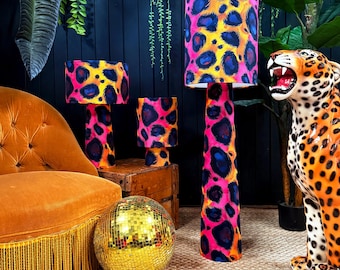 May include: A collection of lamps with vibrant leopard print shades in pink, yellow, and blue. The lamps are of varying sizes and sit on a wooden surface. A gold disco ball and a decorative jaguar statue are also present.