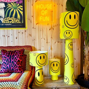 May include: A collection of yellow lamps with smiley faces. The lampshades and bases are yellow with black smiley face graphics. The background includes a neon sign that says "HOUSE OF FUN" and a colorful pillow.