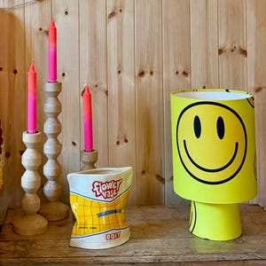 May include: A decorative arrangement featuring a yellow smiley face lamp, three wooden candle holders with pink and orange candles, and a ceramic flower vase. The vase is shaped like a bag of chips with the words "Flower Vase" and "DOIY" printed on it.