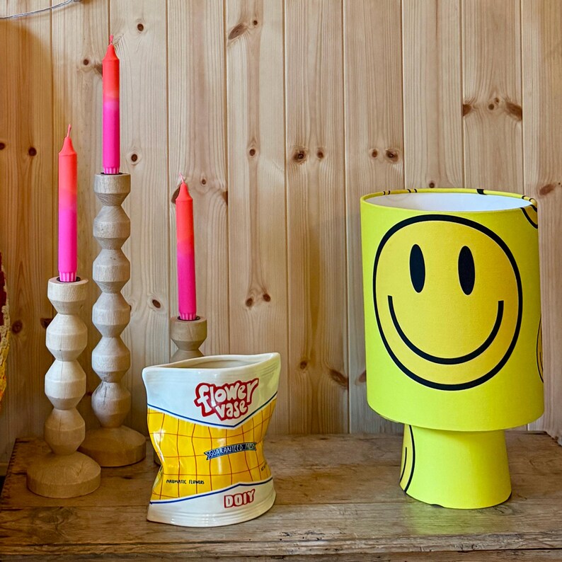 May include: A decorative arrangement featuring a yellow smiley face lamp, three wooden candle holders with pink and orange candles, and a ceramic flower vase. The vase is shaped like a bag of chips with the words "Flower Vase" and "DOIY" printed on it.