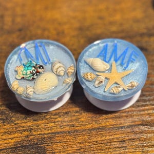 Op de afbeelding: Twee ronde, blauw-witte telefoonhouders met een strandthema. Eén heeft een schildpad en de andere een zeester, omringd door schelpen. De houders hebben de initialen "AMI" en "AMF" in blauw. De achtergrond is een houten oppervlak.