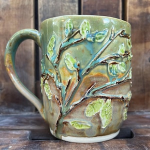 May include: A ceramic mug with a nature-inspired design. The mug has a green and brown glaze with raised leaf and branch details. The handle matches the mug's colour. The mug sits on a white base.