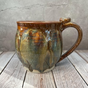 May include: A handmade ceramic mug with a distinctive, textured glaze. The mug has a rounded body and a strong handle. The glaze shifts from brown at the top to green and gold, with white speckles. The mug rests on a white base.