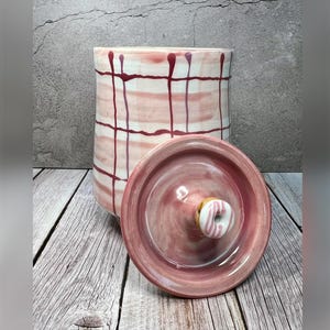 May include: A pink and white ceramic jar with a lid. The jar has a plaid pattern with red and white stripes. The lid has a pink and white swirl design.