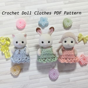 May include: Image of three animal figurines wearing handmade crochet dresses. The figurines are a cat, a rabbit, and a lamb. The dresses are blue, green, and pink. The text "Crochet Doll Clothes PDF Pattern" is at the top.