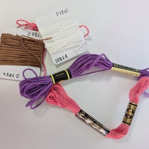 May include: A collection of embroidery floss in various colors, including brown, white, purple, and pink. The floss is organized with labels marked with numbers. The DMC brand floss is visible, with the number 25 on the label.