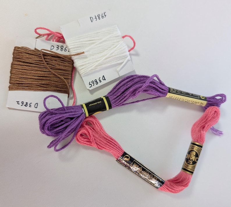 May include: A collection of embroidery floss in various colors, including brown, white, purple, and pink. The floss is organized with labels marked with numbers. The DMC brand floss is visible, with the number 25 on the label.