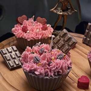 Könnte beinhalten: Zwei rosa und graue Cupcake-Kerzen mit Schokoladenakzenten. Die Kerzen sind mit Streuseln und Schokoladenstückchen verziert.