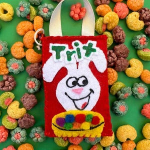 May include: A red felt bag with a white bunny face and the word "Trix" in green. The bag is filled with colorful cereal pieces. A yellow ribbon handle is attached to the bag. The background is green.