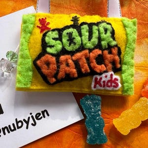 May include: A felt Sour Patch Kids candy wrapper with green and orange lettering, a red candy, and three gummy candies. The wrapper is yellow with green trim and a white ribbon. A white card with text is in the background.