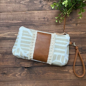 May include: A beige and white patterned clutch with a brown leather accent and a fringe trim. The clutch has a brown leather strap.